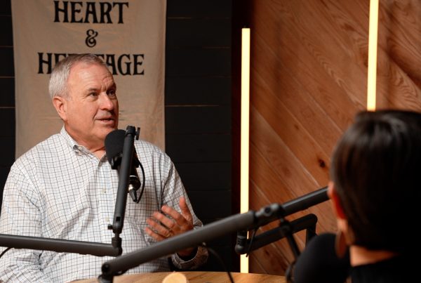 A man in a podcast room, decorated with warm wood tones, LED tube lighting and a flag that reads Heart and Heritage, with a mic in front of his face while he continues talking.