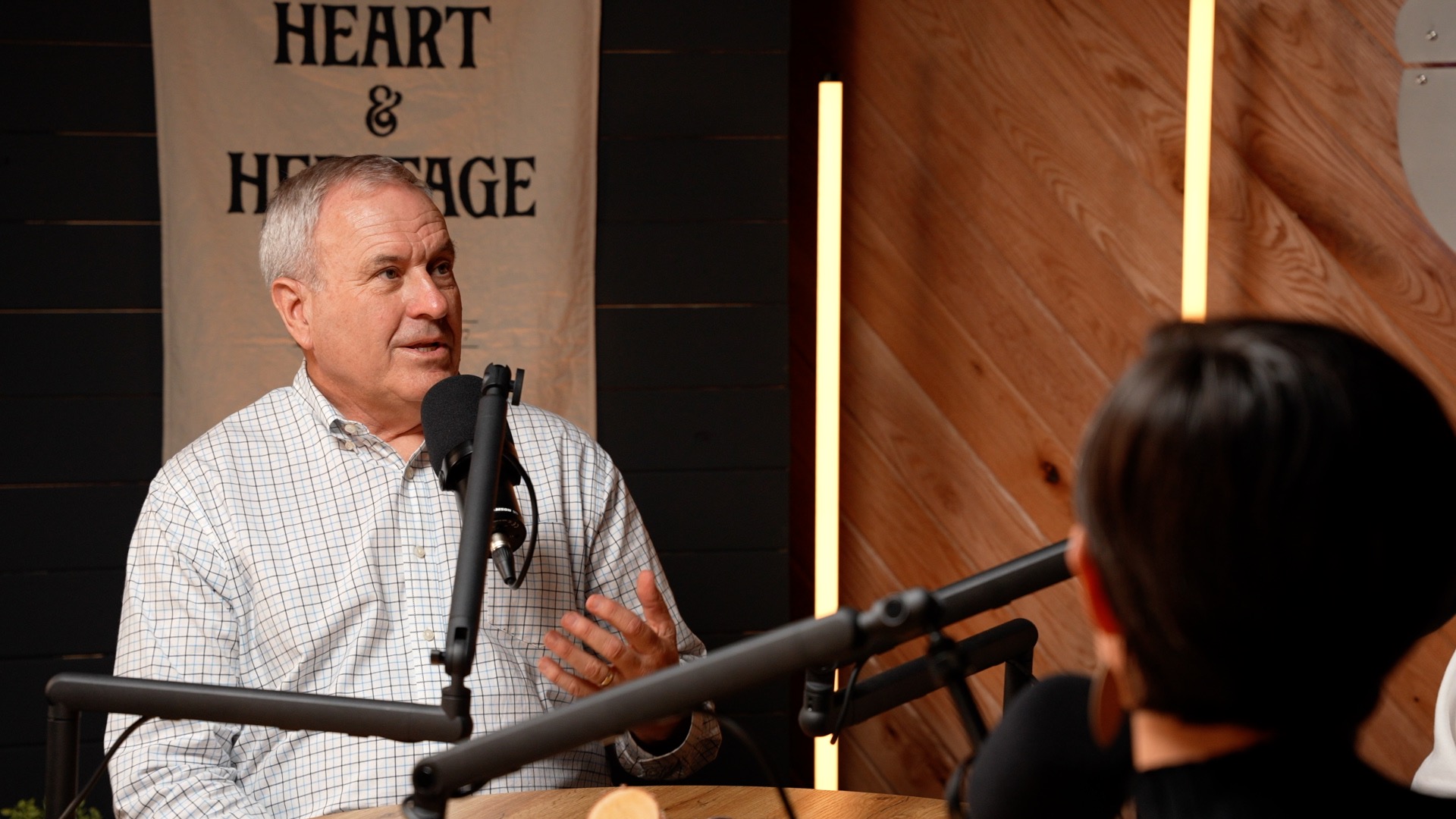 A man in a podcast room, decorated with warm wood tones, LED tube lighting and a flag that reads Heart and Heritage, with a mic in front of his face while he continues talking.