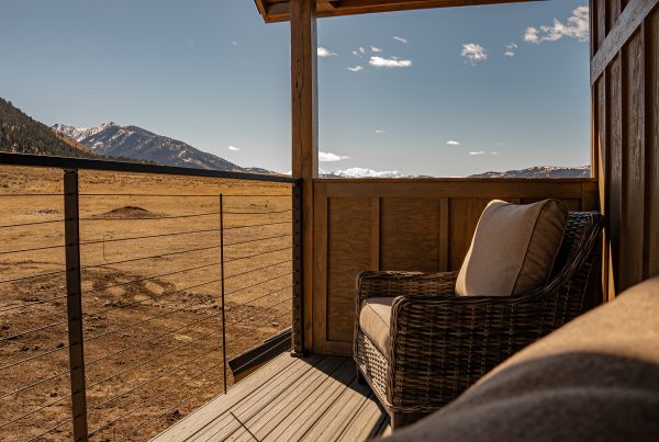 A view from a covered wooden balcony or deck looking out over a scenic, expansive landscape. A comfortable wicker armchair with light tan cushions sits in the corner of the deck, tucked against vertical wood-paneled siding. A modern cable railing encloses the deck, offering an unobstructed view of a vast, dry, rolling valley floor that leads to majestic, snow-capped mountains under a bright blue sky with scattered light clouds.
