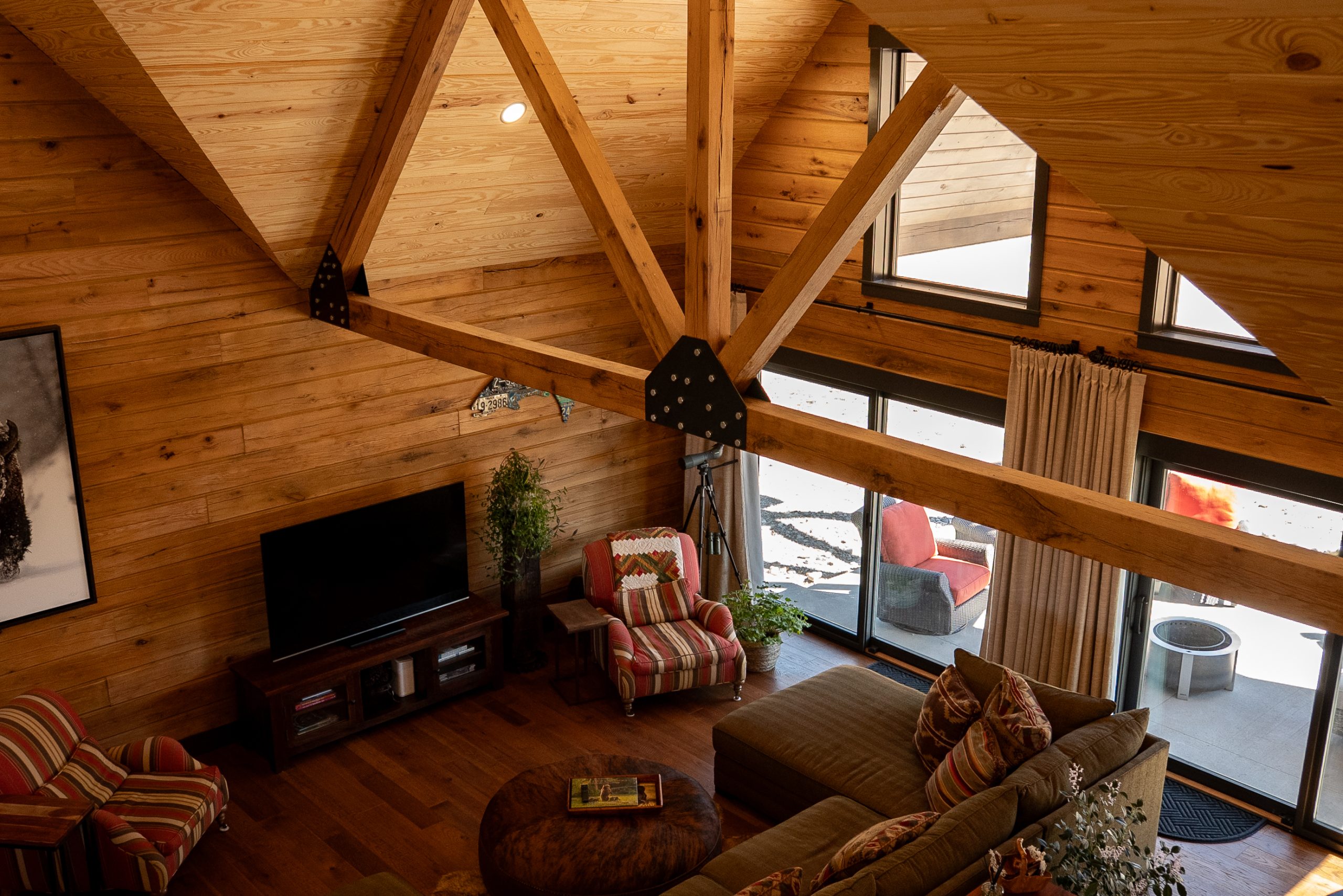 A high-angle, overhead view of a rustic living room inside a log home, featuring soaring vaulted ceilings lined with warm wood paneling and highlighted by a massive timber king post truss with black metal plates. Below, the cozy sitting area includes a green sectional sofa with patterned throw pillows, a round tufted leather ottoman, a television on a wooden media console, and two red-and-tan striped armchairs. Large sliding glass doors and high clerestory windows flood the space with natural light and look out onto a concrete patio with outdoor seating and a fire pit.