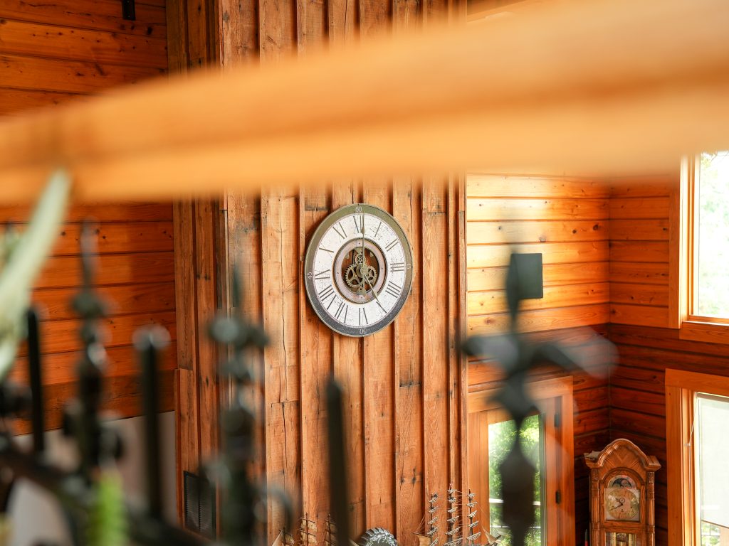 A close-up interior view looking across a wooden railing at a prominent decorative wall clock mounted on a vertical wood-paneled wall. The round clock features Roman numerals and a central cutout that displays exposed brass gears. The surrounding walls are made of warm, horizontal wood logs, and a classic wooden grandfather clock is partially visible in the lower right corner. The foreground is slightly blurred, showing the dark metal tips of a wrought-iron railing and a wooden beam.