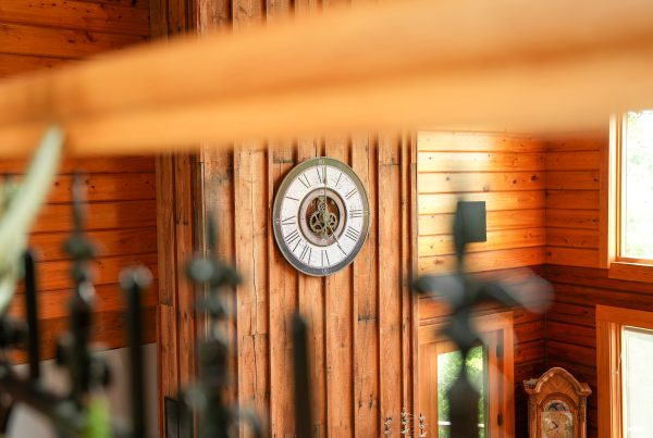 A close-up interior view looking across a wooden railing at a prominent decorative wall clock mounted on a vertical wood-paneled wall. The round clock features Roman numerals and a central cutout that displays exposed brass gears. The surrounding walls are made of warm, horizontal wood logs, and a classic wooden grandfather clock is partially visible in the lower right corner. The foreground is slightly blurred, showing the dark metal tips of a wrought-iron railing and a wooden beam.