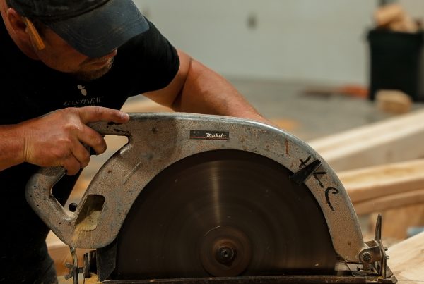 Craftsman cutting a large timber beam with a circular saw in a woodworking shop.