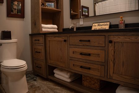A close-up, low-angle view of a bathroom vanity and toilet in a rustic home. The large oak vanity features black hardware, multiple drawers, a dark stone countertop, and an open bottom shelf holding folded white towels and a woven storage basket. To the left sits a white toilet. On the countertop, a single silver faucet is visible next to a decorative sign reading "the Mountains are calling." Built-in wooden shelving above the counter holds stacked towels and framed pictures, with additional nature-themed framed art hanging on the neutral-toned wall.