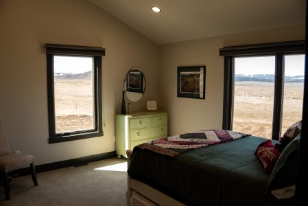 A well-lit bedroom featuring a vaulted ceiling and neutral-toned drywall, contrasting with dark window trim and baseboards. In the foreground, a bed is styled with a dark green comforter and a patterned, geometric-print throw blanket across the foot. Nestled between two large windows that offer sweeping views of an open, arid prairie landscape is a light green vintage-style dresser with a round mirror hanging above it. A simple upholstered accent chair sits on the carpeted floor to the far left.