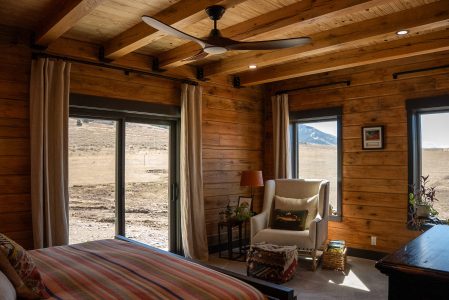A cozy, sunlit corner of a rustic log home bedroom featuring warm, horizontal wood-paneled walls and exposed timber ceiling beams with a modern wooden ceiling fan. In the corner, an off-white wingback armchair sits next to a patterned ottoman pouf, a small side table with a lamp, and a basket of books. Two large windows and a sliding glass door with light-colored curtains frame views of a vast, sunny, rolling hillside landscape under a clear sky. A portion of a bed with a colorful, warm-toned striped duvet is visible in the foreground on the left.