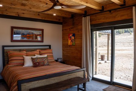 A rustic bedroom interior featuring a large bed with a warm, multi-colored striped duvet and matching pillows in shades of orange, brown, and green. The room showcases exposed wooden ceiling beams with a modern, three-blade wooden ceiling fan, an accent wall with horizontal wood paneling, and a painted drywall accent wall behind the bed. To the right, a large sliding glass door framed by light-colored curtains opens up to an outdoor patio overlooking a vast, rolling landscape of dry grass and distant hills. Framed artwork, including a scenic mountain landscape above the bed and a small, colorful painting on the wood wall, adds a decorative touch.