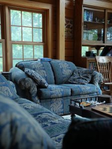 A cozy sitting area inside a log home featuring patterned blue and cream sofas, a wooden coffee table with a chessboard, large paneled windows, and a built-in wooden bookcase filled with books.