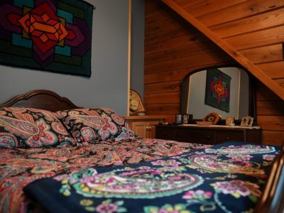 A cabin bedroom featuring a bed with a colorful paisley comforter, a dark wood dresser with an arched mirror, blue walls, a geometric tapestry, and a slanted wood-paneled ceiling accentuating the cabin style.