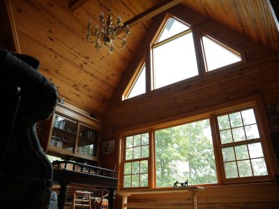 A low-angle view of a log cabin living room showcasing tall A-frame windows, a brass chandelier hanging from a vaulted tongue-and-groove wood ceiling, and a large window looking out onto green trees.