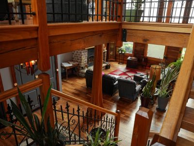 A high-angle interior view looking down from a wooden staircase into a spacious, multi-level living room. The home features thick timber support beams, polished hardwood floors, and wooden railings with ornate black metal balusters that wrap around an upper loft area. Below, the main living space is furnished with dark leather seating, a gray armchair, potted snake plants, and a vibrant red patterned area rug. In the background, a brick fireplace with a wood-burning stove stands out against horizontal wood log walls.