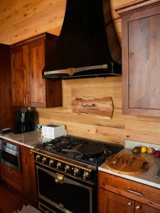 A rustic kitchen featuring a large black and brass gas range cooker with matching hood, warm wood paneled walls, and custom wooden cabinetry. A live-edge wood sign reading "Together is my favorite place to be" hangs on the backsplash above the stove.