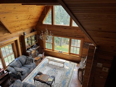 High-angle view of a cozy log cabin living room featuring cathedral wood ceilings, large windows overlooking a forest, patterned blue sofas, an ornate area rug, and a brass chandelier.